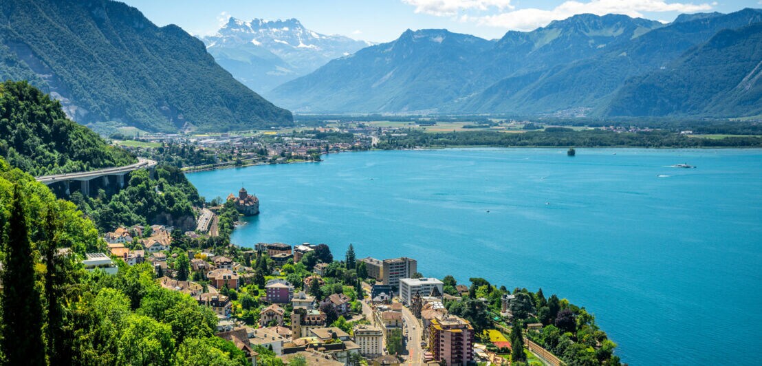 Blick auf den Genfersee mit der Stadt Montreux, umgeben von grünen Bergen und schneebedeckten Alpen im Hintergrund