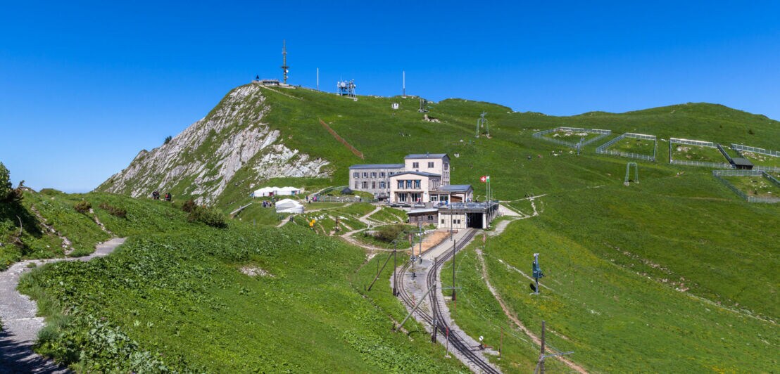 Grüner Berg mit Wanderweg, Bergbahn und Gebäude unter klarem blauem Himmel