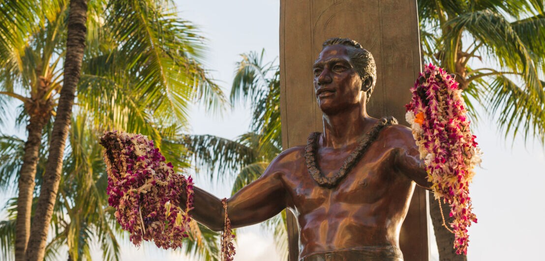 Bronzestatue von Surf-Legende Duke Kahanamoku mit ausgebreiteten Armen, an denen Blumenketten hängen.