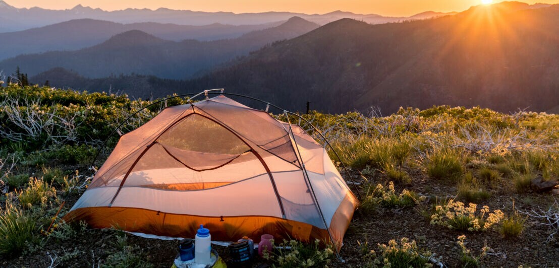 Zelt auf Berggipfel bei Sonnenaufgang mit Blick auf mehrere Bergketten und Campingausrüstung im Vordergrund