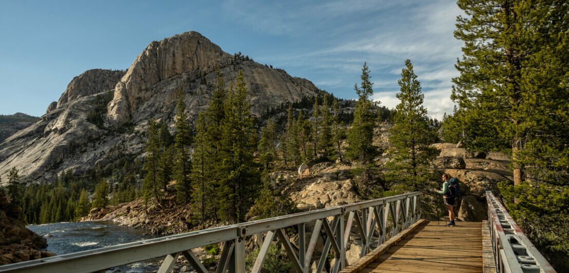 Wanderer mit Rucksack auf einer Holzbrücke über einen Fluss, umgeben von Kiefern und Bergen bei bewölktem Himmel