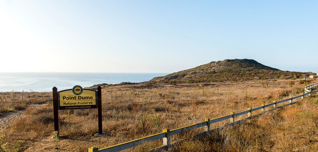 Schild mit der Aufschrift Point Dume Natural Preserve vor trockenem Grasland und einem Hügel, im Hintergrund das Meer.