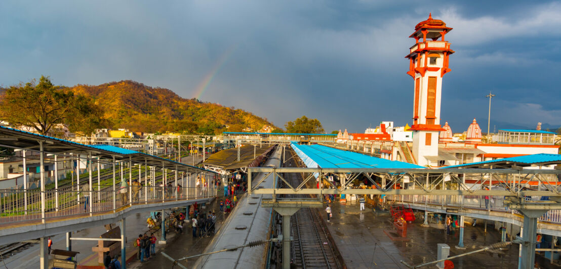 Blick auf einen Bahnhof mit Bahnsteigen und einem Zug, im Hintergrund ein hoher Turm und bewaldete Hügel unter bewölktem Himmel