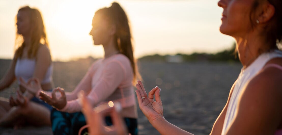 Drei Frauen sitzen im Schneidersitz am Strand und meditieren bei Sonnenuntergang
