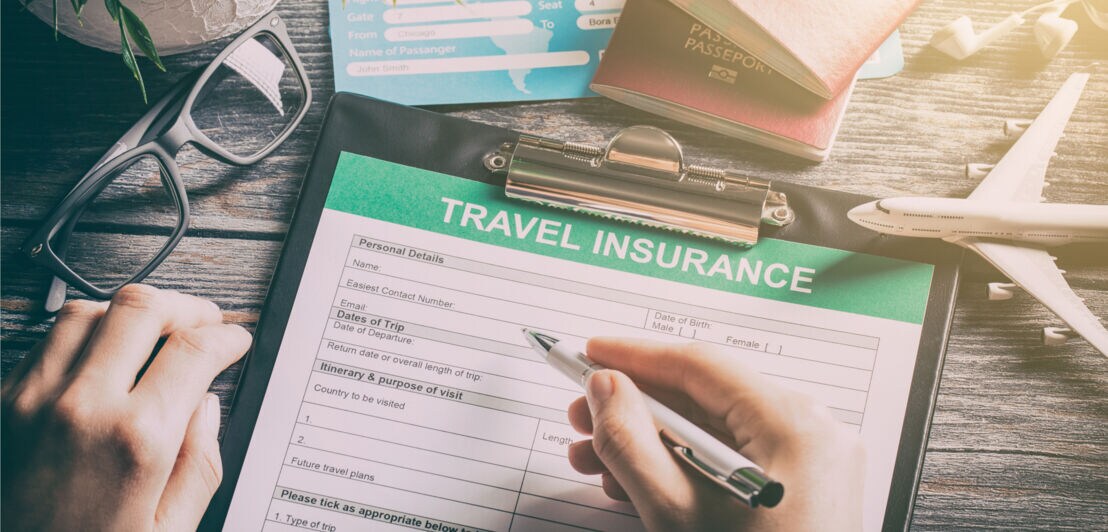 Person füllt ein Formular zur Reiseversicherung auf einem Klemmbrett aus, umgeben von Reisepässen, Flugtickets, einer Brille und einem Modellflugzeug