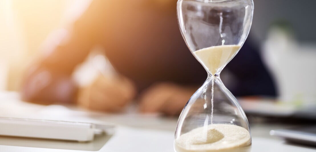 Sanduhr auf einem Schreibtisch mit verschwommenem Hintergrund einer Person, die den Kopf auf den Tisch legt