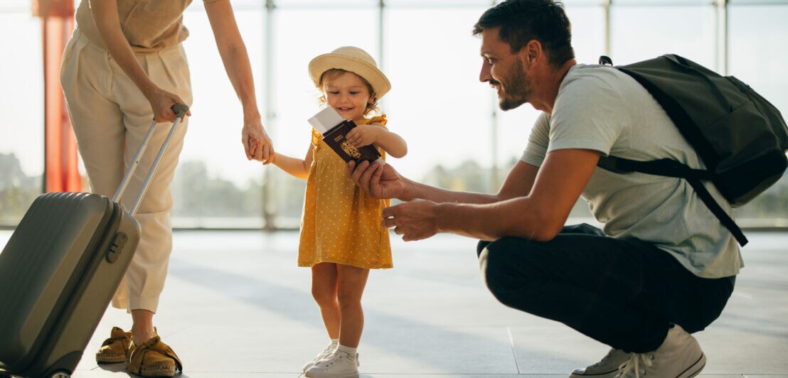 Mutter mit Rollkoffer hält die Hand eines kleinen Mädchens in gelbem Kleid und Sonnenhut, Vater kniet und zeigt Reisepässe