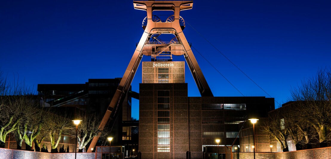 Fördergerüst der Zeche Zollverein mit beleuchtetem Zollverein-Schriftzug bei Abenddämmerung.
