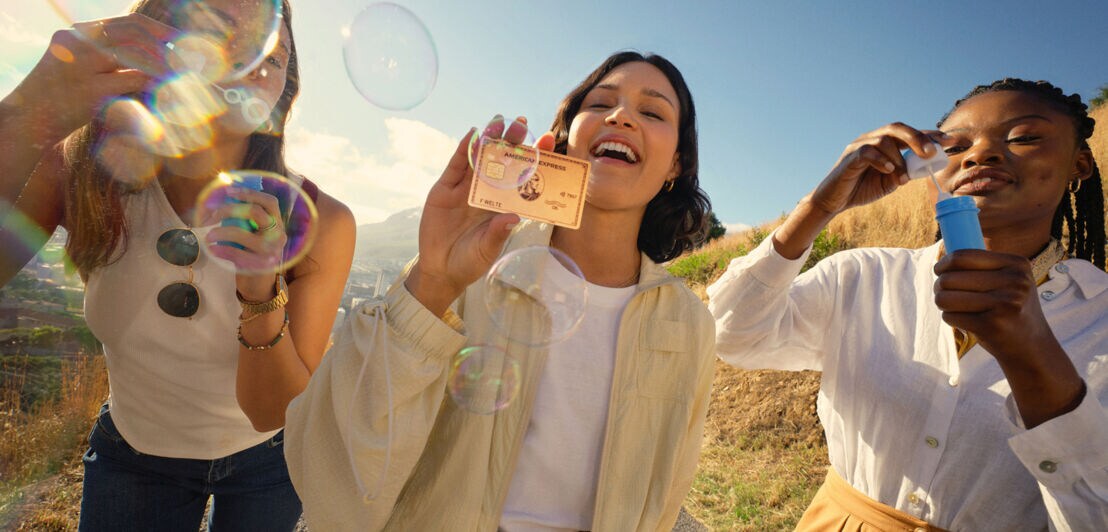 Drei Frauen unter freiem Himmel bei Sonnenschein, von denen zwei Seifenblasen blasen und eine die roséfarbene Kreditkarte von American Express präsentiert.