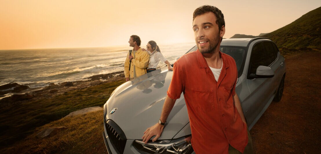 Ein junger Mann im roten Hemd lehnt an der Motorhaube eines BMWs, dahinter zwei weitere Personen an einer Meeresküste bei Sonnenuntergang.