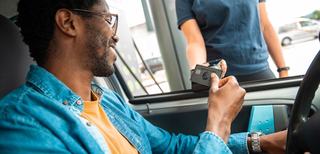 Ein Mann im Jeanshemd am Steuer eines Autos bezahlt kontaktlos mit einer silbernen American Express Kreditkarte an einem Kartenlesegerät, das eine Person in das geöffnete Autofenster hält.
