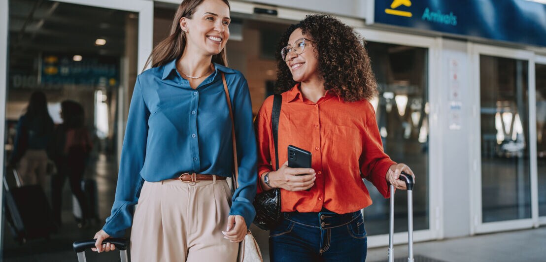 Zwei lachende, elegant gekleidete Frauen mit Trolleys beim Verlassen eines Flughafenterminals.
