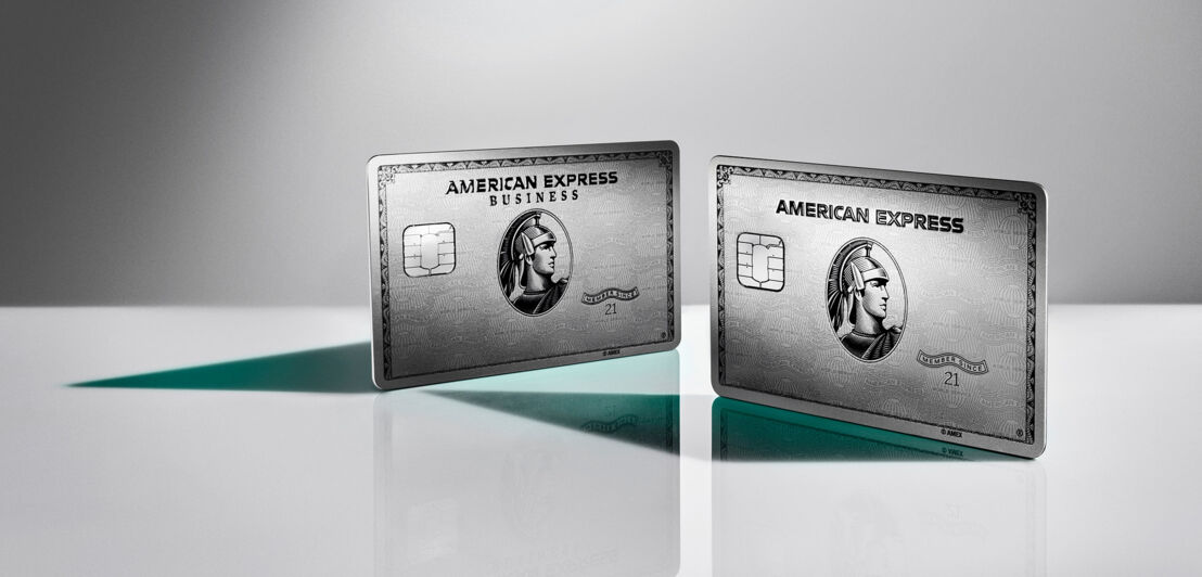 Studioaufnahme einer aufgestellten silbernen American Express Business Kreditkarte neben einer aufgestellten silbernen American Express Kreditkarte auf einer weißen Spiegelfläche vor grauem Hintergrund.