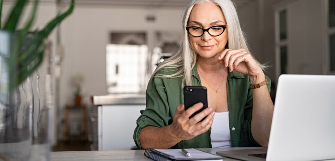 Eine Frau mit langen weißen Haaren sitzt vor einem Laptop an einem Schreibtisch im Büro und schaut zufrieden auf ein Smartphone in ihrer Hand.