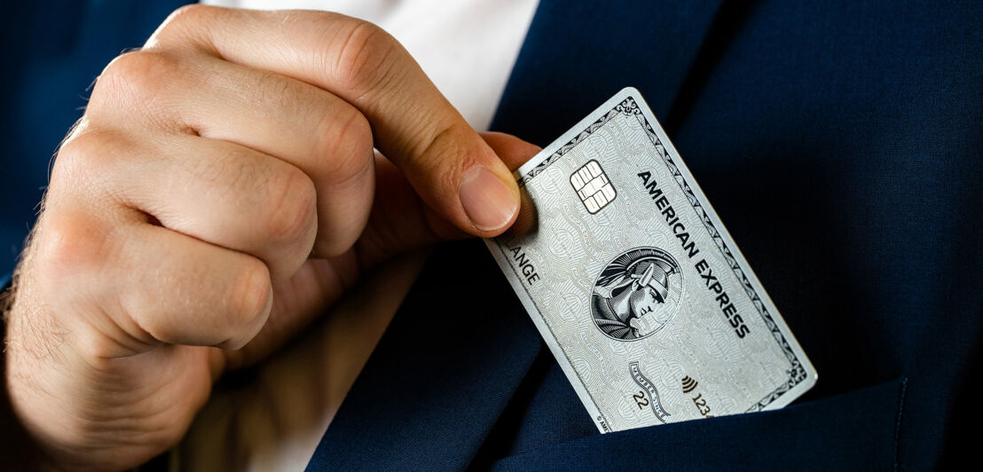 Nahaufnahme einer männlichen Hand, die eine silberne Kreditkarte von American Express aus der Brusttasche eines blauen Jacketts zieht.