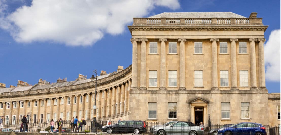 Historisches Gebäude mit Säulen und vielen Fenstern, davor parkende Autos und Menschen auf dem Bürgersteig
