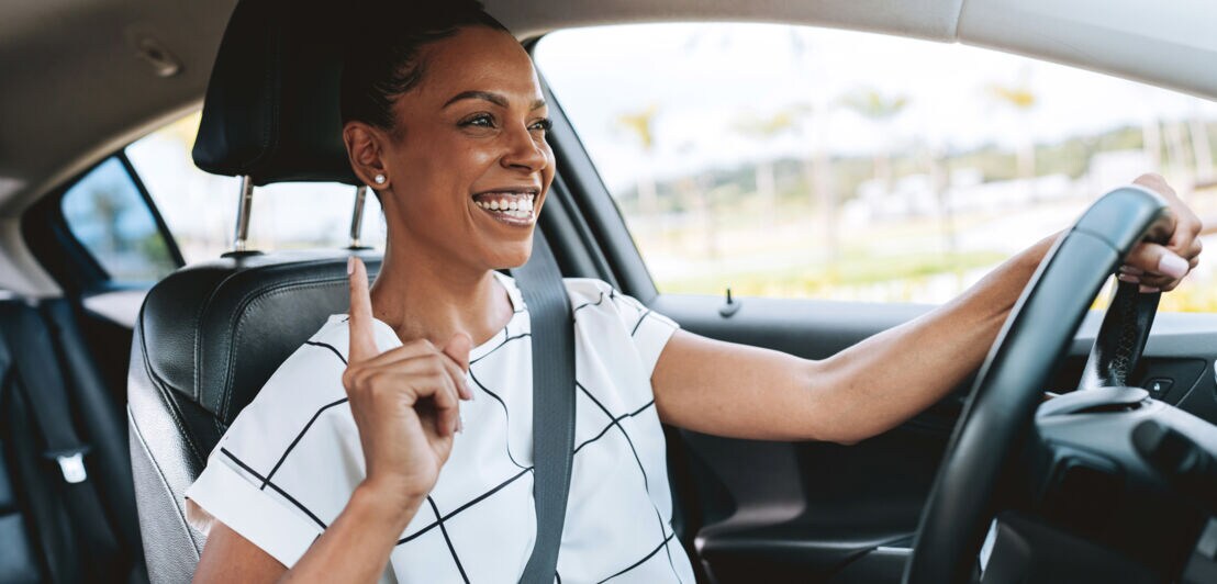 Eine lachende Frau im weißen Shirt am Steuer eines Autos mit einer Hand am Lenkrad.