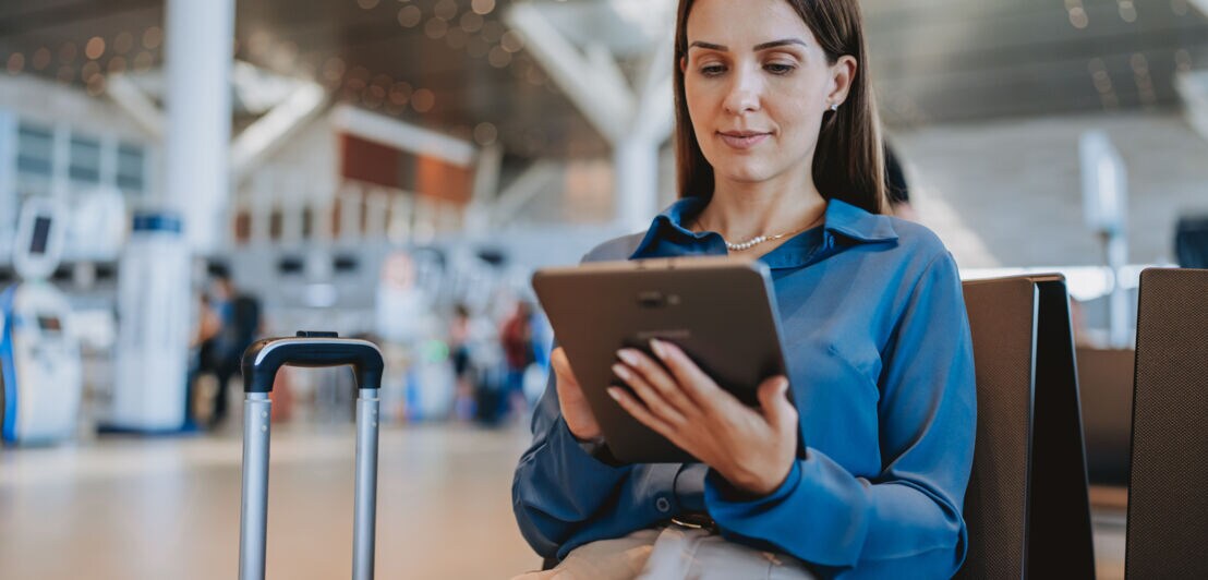 Eine elegant gekleidete Frau sitzt im Wartebereich eines Flughafenterminals und bedient ein Tablet, neben ihr steht ein Trolley.