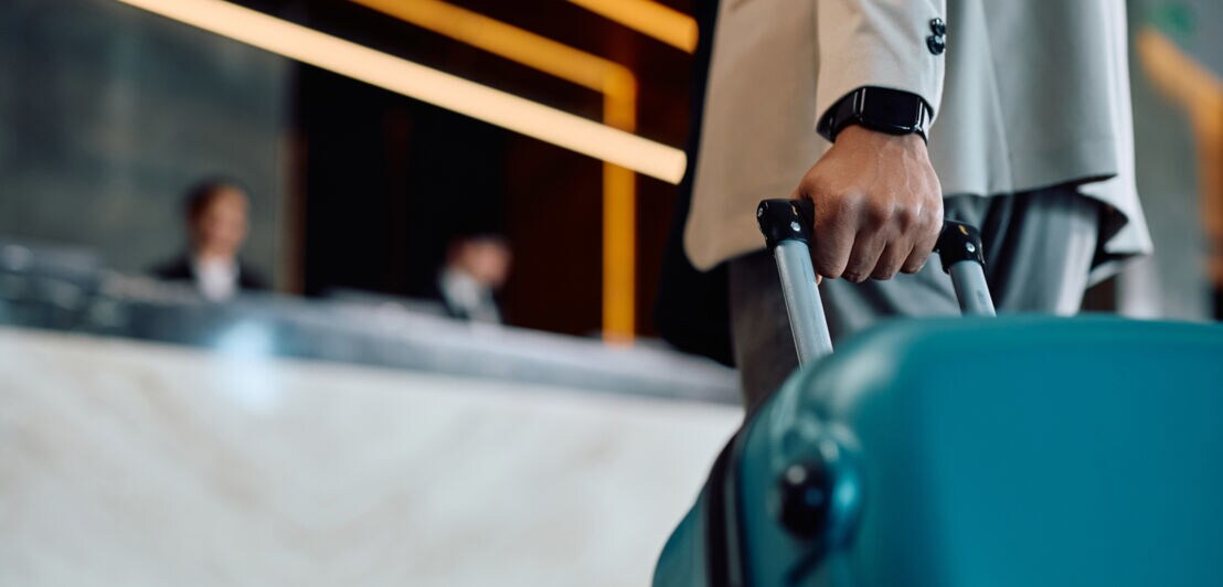 Nahaufnahme einer Hand am Griff eines türkisfarbenen Hartschalenkoffers, dahinter eine Hotelrezeption in Unschärfe.