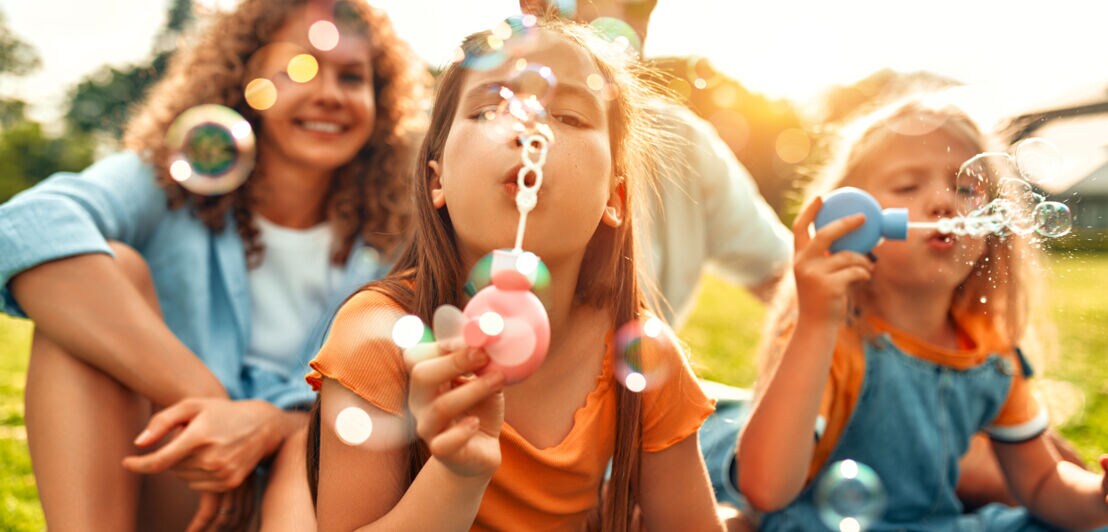 Vier Personen, davon zwei Kinder, sitzen draußen auf Rasen und pusten Seifenblasen.