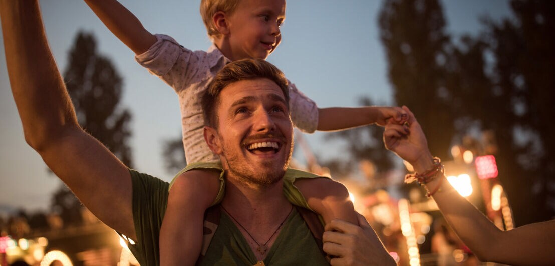 Ein Mann im Dunkeln mit kleinem Kind auf den Schultern, im Hintergrund Bäume und Beleuchtung.