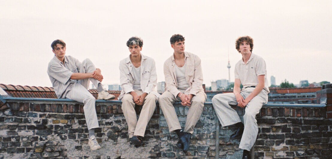 Vier junge Männer, die Mitflieder der Band Oswald, in heller Freizeitkleidung sitzen auf einer Mauer mit Stadtblick im Hintergrund.