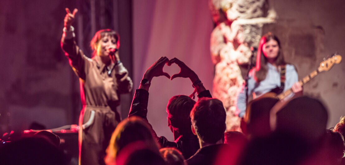 Publikum bei einem Konzert formt mit den Händen ein Herz, während zwei Musikerinnen auf der Bühne spielen