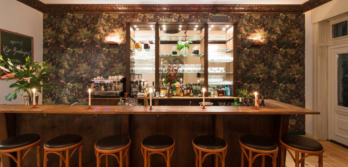 Holztresen mit sieben Barhockern vor einer Wand mit floralem Muster und beleuchtetem Regal mit Gläsern und Flaschen