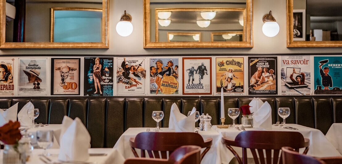 Restaurant mit weißen Tischdecken, gefalteten Servietten, Weingläsern und Stühlen vor einer Wand mit goldgerahmten Spiegeln und Vintage-Filmplakaten