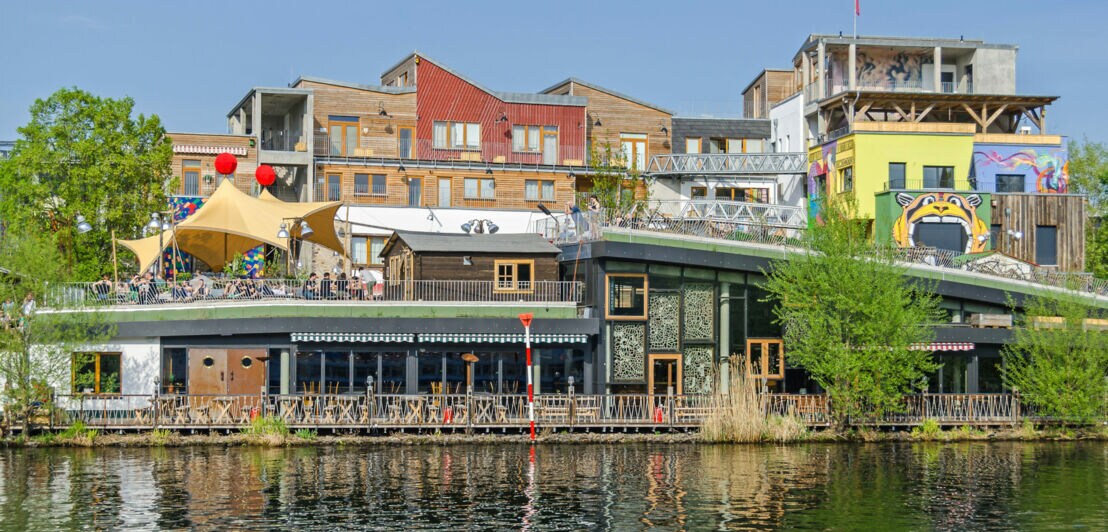 Bunte Gebäude und Cafés am Ufer eines Kanals mit Menschen auf der Terrasse und Spiegelung im Wasser