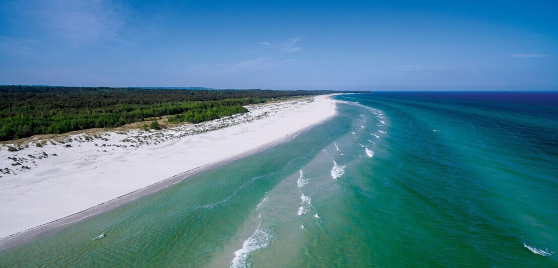 Luftaufnahme eines langen weißen Sandstrands mit grünem Wald im Hintergrund und ruhigem Meer unter blauem Himmel