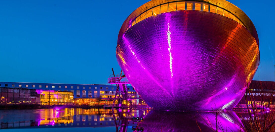 Wissenschaftsmuseum Universum Bremen in Form einer silbernen Schale, beleuchtet in Lila und Orange, am Wasser bei Nacht.