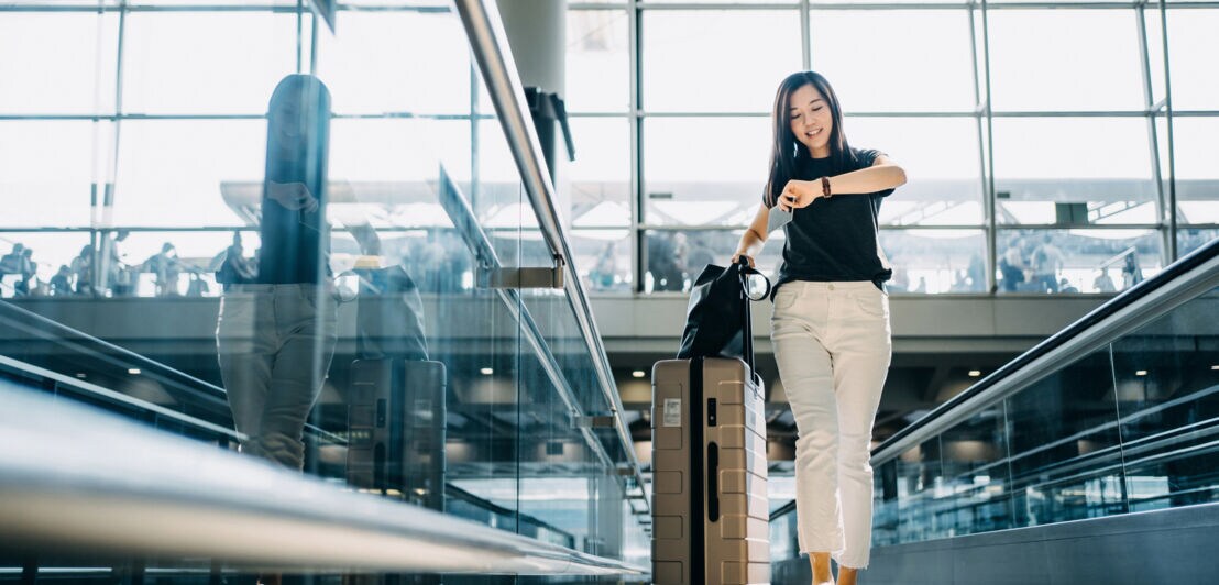 Eine Frau mit Rollkoffer schaut auf Ihre Armbanduhr in einem Flughafenterminal.