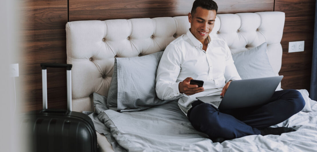 Mann sitzt in Hemd und Anzughose im Schneidersitz auf einem Hotelbett und telefoniert, auf dem Schoß einen Laptop.