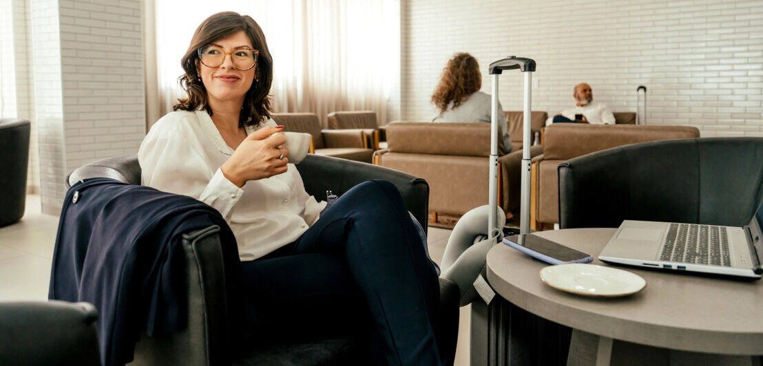 Frau sitzt in exklusiver Lounge, in der Hand eine Kaffeetasse, vor sich ein kleiner Tisch mit Laptop und Smartphone.