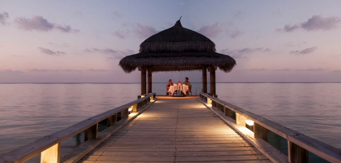 Ein Paar beim Candle-Light-Dinner unter einem Pavillon am Ende eines Stegs am Meer.