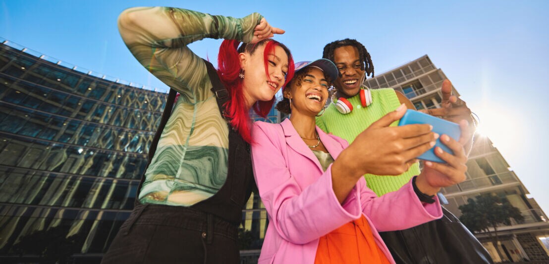 Drei junge, stylische Personen in bunter Kleidung schauen gut gelaunt gemeinsam auf ein Smartphone in urbaner Umgebung unter blauem Himmel.