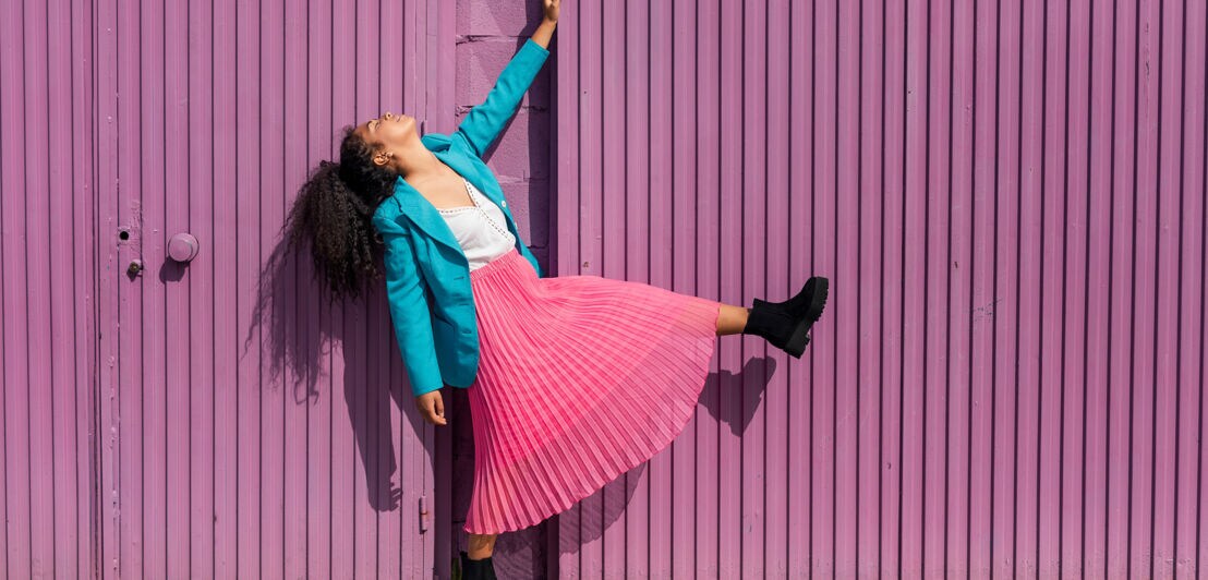 Eine Frau im türkisblauen Balzer und rosafarben Plisseerock posiert vor einer lilafarbenen Fassade.