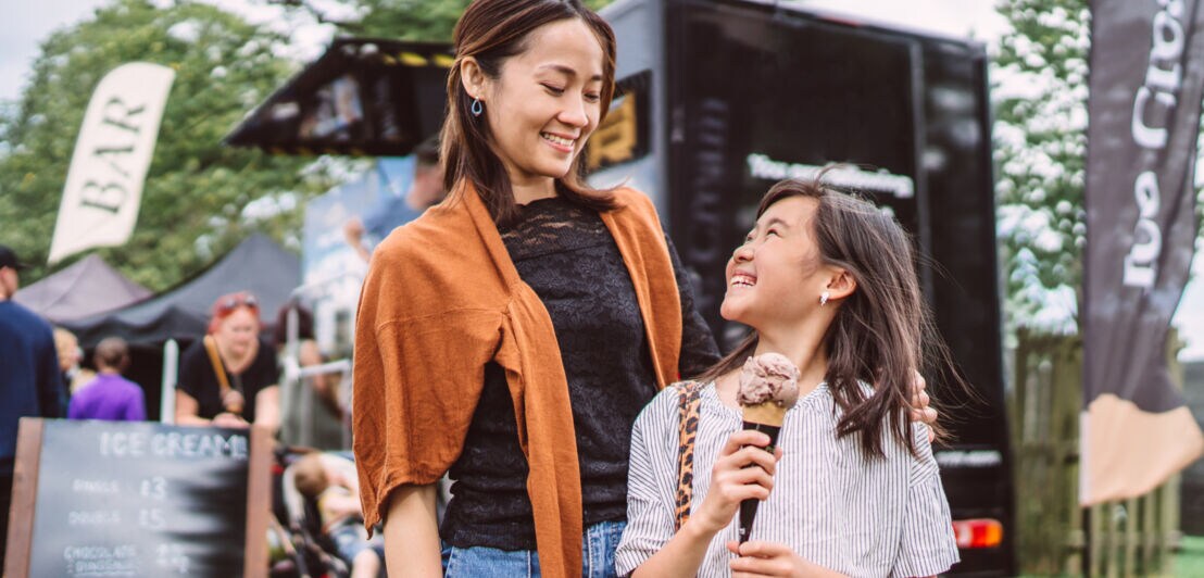 Frau mit orangefarbenem Pullover um die Schultern und Kind mit Eiscreme in der Hand auf einem Festival mit Foodtrucks im Hintergrund.