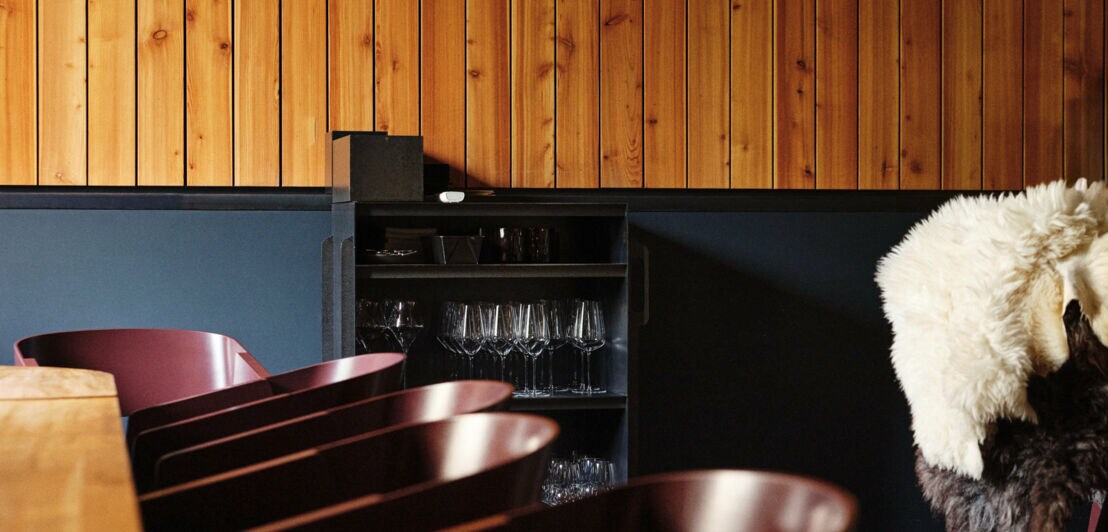 Knapper Ausschnitt des Gastraums mit dunklen Sesseln am langen Tisch und einem kleinen Schrank mit Gläsern, der an der halb holzvertäfelten und halb petrolfarben gestrichenen Wand steht.