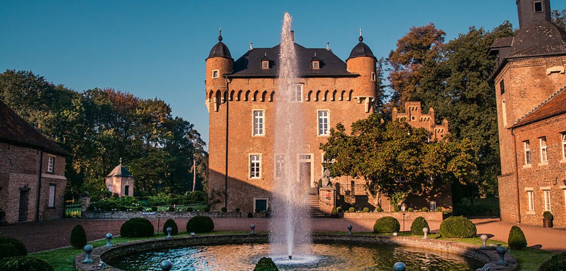 Brunnen mit Fontäne vor einem historischen Schloss mit runden Türmen und Zinnen bei Sonnenuntergang.