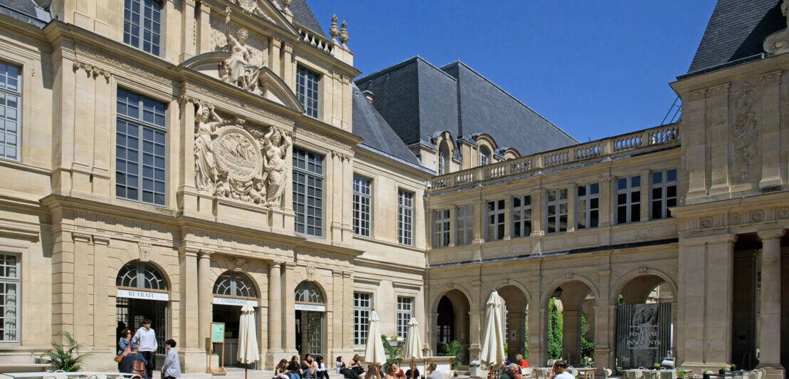 Innenhof eines historischen Gebäudes mit Arkaden, Sitzgruppen und Menschen, die bei Sonnenschein entspannen