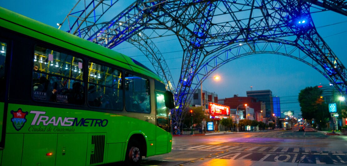 Grüner Transmetro-Bus unter beleuchtetem Metallbogen in der Stadt Guatemala bei Dämmerung