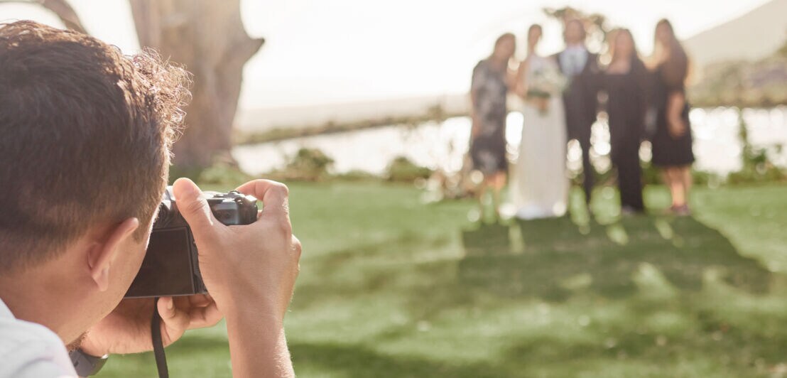 Fotograf fotografiert Brautpaar und Gäste bei Hochzeit im Freien mit See im Hintergrund.