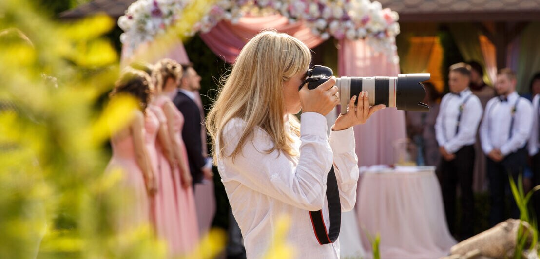Frau mit langen blonden Haaren fotografiert mit einer Kamera mit Teleobjektiv bei einer Hochzeit im Freien.