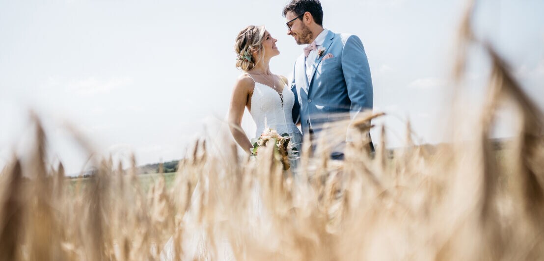 Braut in weißem Kleid mit Blumenstrauß und Bräutigam im blauen Anzug stehen in einem Weizenfeld.