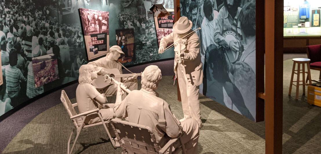 Drei weiße Skulpturen spielen Bluegrass-Musikinstrumente in einem Museum mit historischen Fotos an den Wänden