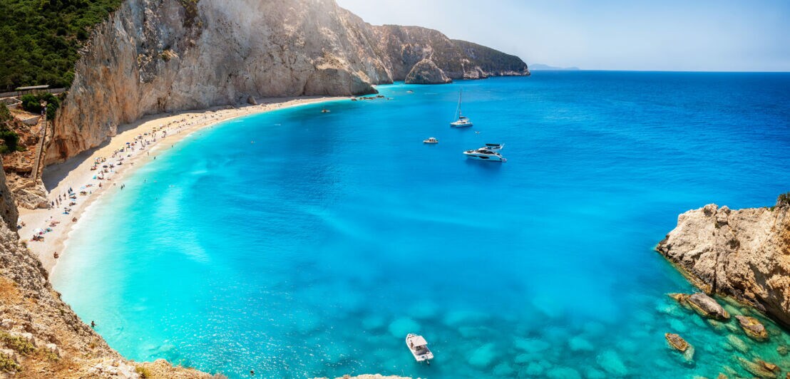 Blick von oben auf einen Sandstrand in einer von Felsen gesäumten Bucht am türkisblauen Meer.