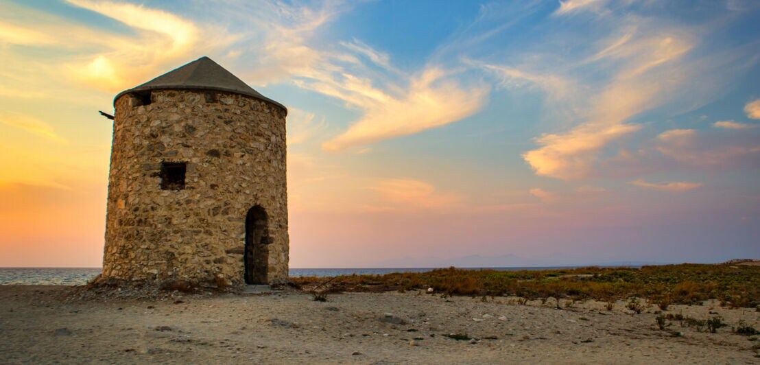 Historische Windmühle aus Stein an einem Strand bei Sonnenuntergang.
