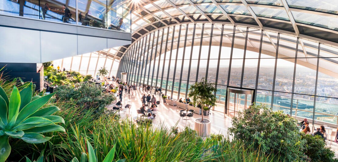 Großer Wintergarten mit Glasdach und Panoramafenstern, viele Pflanzen und Menschen an Tischen, Stadtblick im Hintergrund.